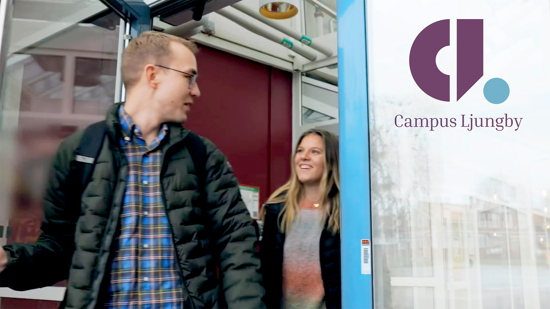En ung man och en ung kvinna går in genom glasdörrarna, med Campus Ljungbys lila logotyp.