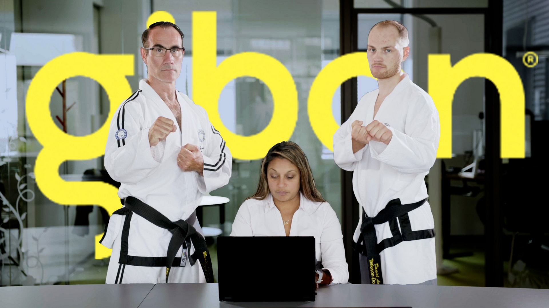 Stillbild ur Gibons reklamfilm: två män i taekwondodräkt står vakt bakom en kvinna vid en laptop, framför Gibons gula logotyp.