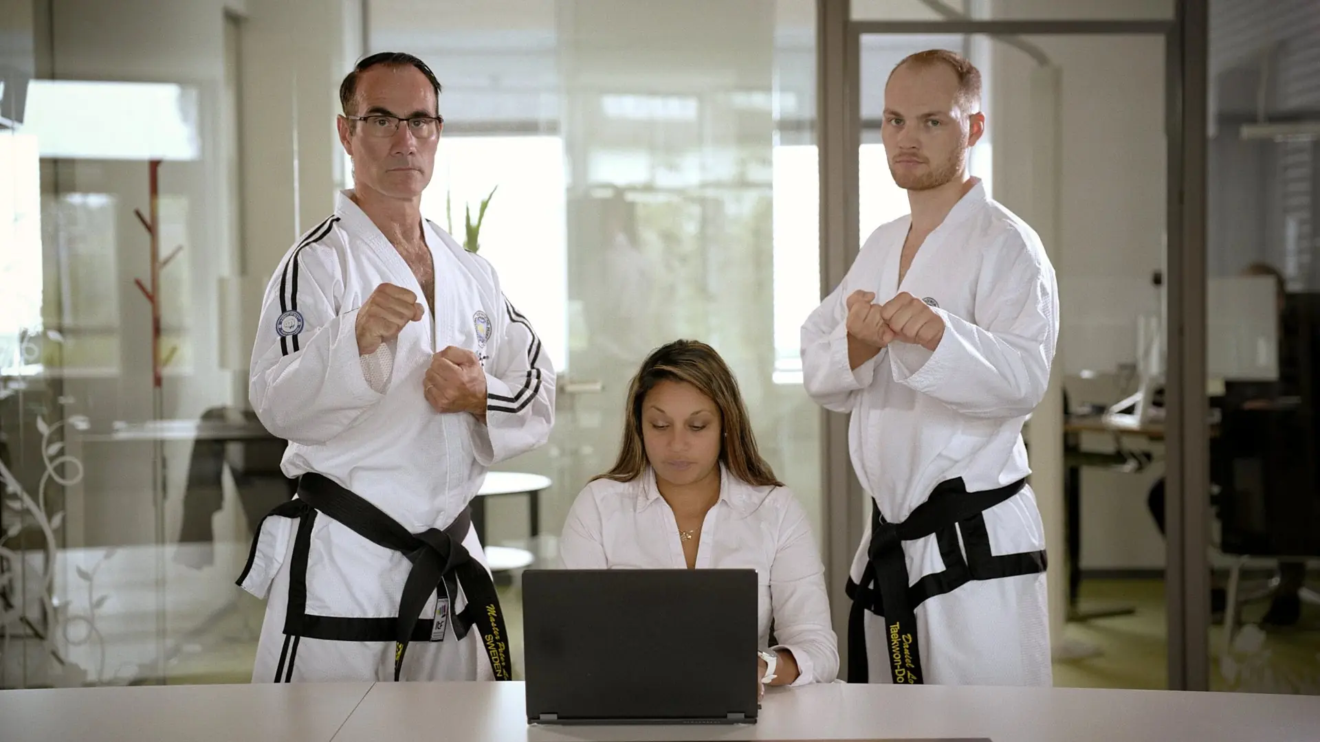 Stillbild ur Gibons reklamfilm: två män i taekwondodräkt står vakt bakom en kvinna vid en laptop, i ett kontor med glasväggar.