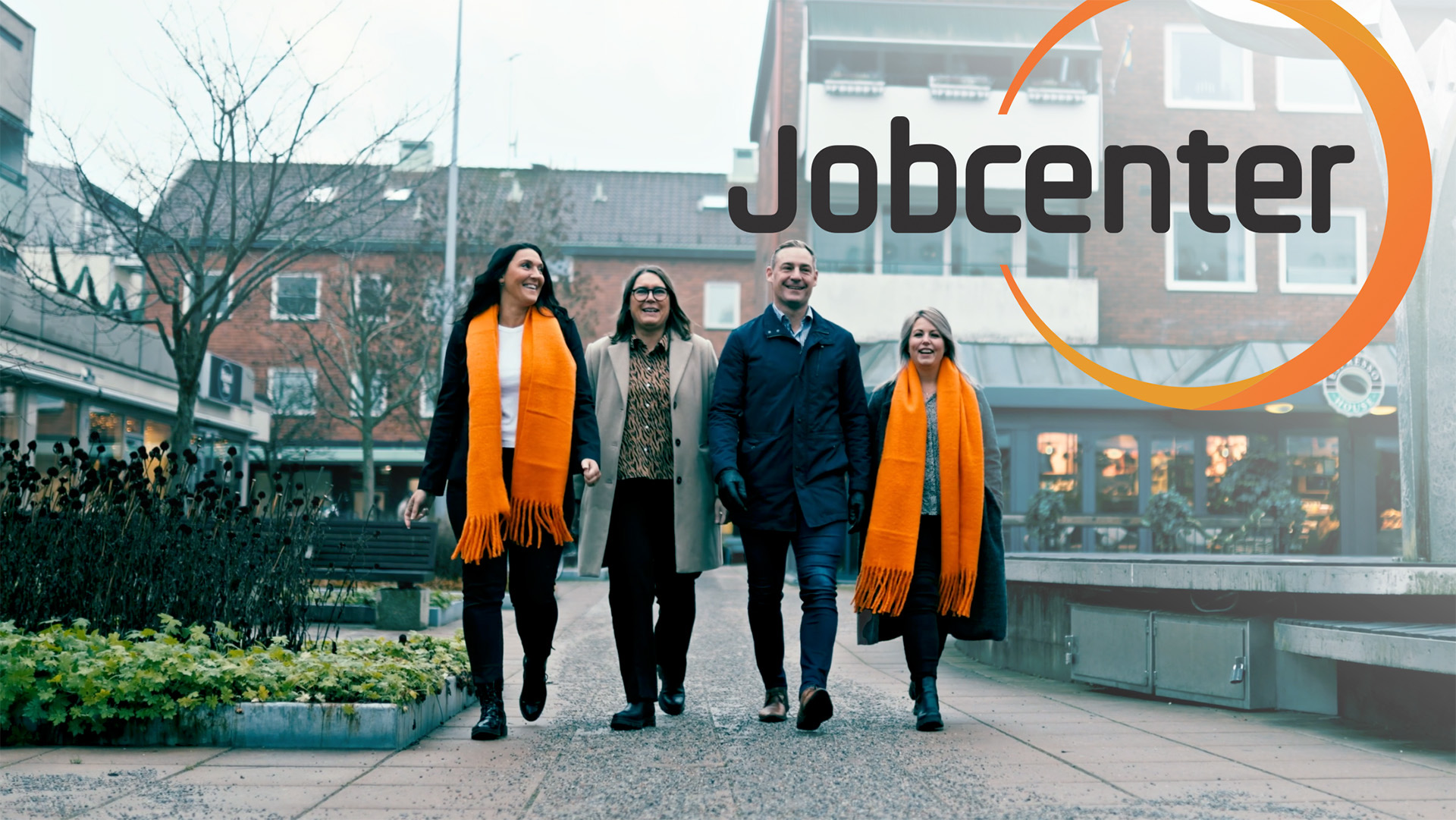 Fyra personer i orange halsdukar går leende över ett torg, med Jobcenters orange logotyp.