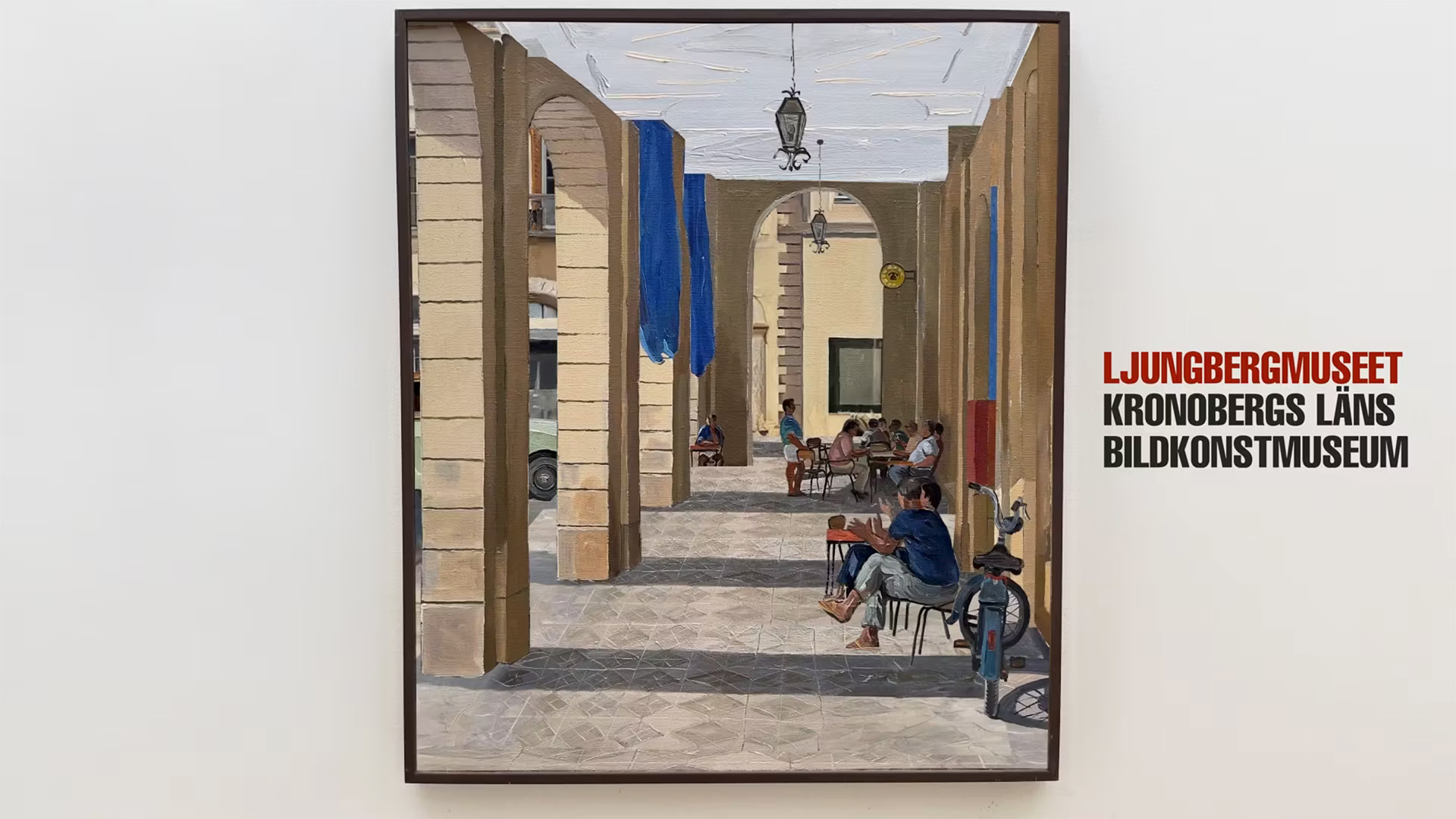 Sven Ljungbergs målning av en arkad med kafébord hänger på museets vägg, bredvid Ljungbergmuseets logotyp med texten Kronobergs läns bildkonstmuseum.