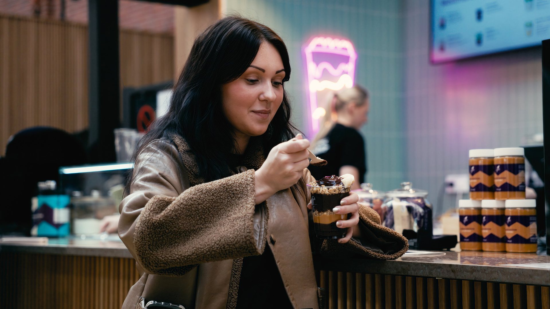 En ung kvinna äter ur en Oakberry-bägare med açaí i lager vid disken. I bakgrunden en rosa neonskylt formad som en bägare och staplade Oakberry-burkar.