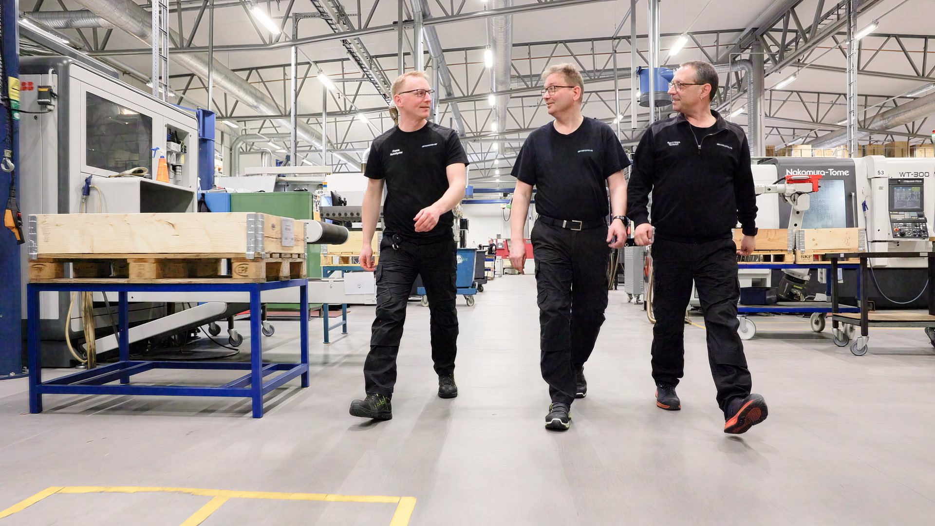 Tre medarbetare i svarta arbetskläder går sida vid sida genom maskinhallen, bland CNC-maskiner och lastpallar, och pratar med varandra.