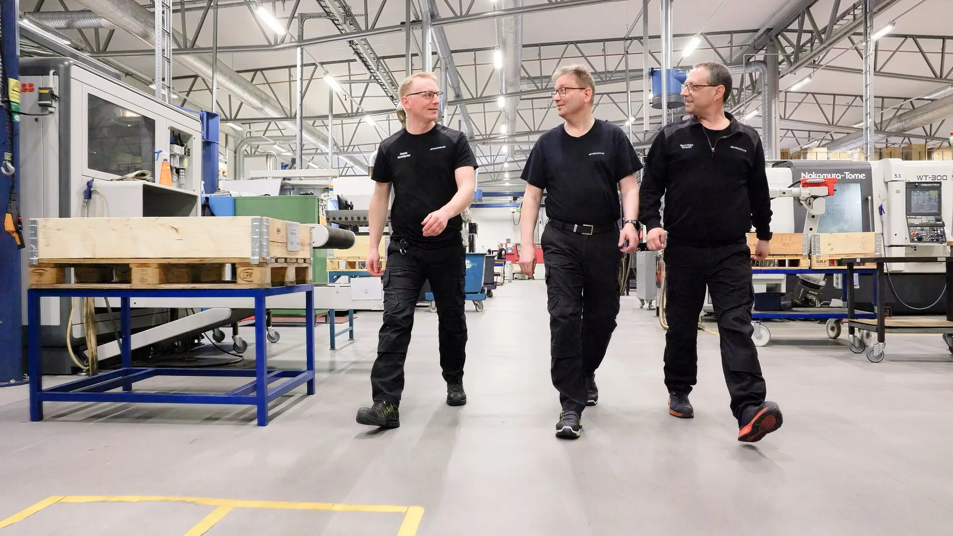 Tre medarbetare i svarta arbetskläder går sida vid sida genom maskinhallen, bland CNC-maskiner och lastpallar, och pratar med varandra.