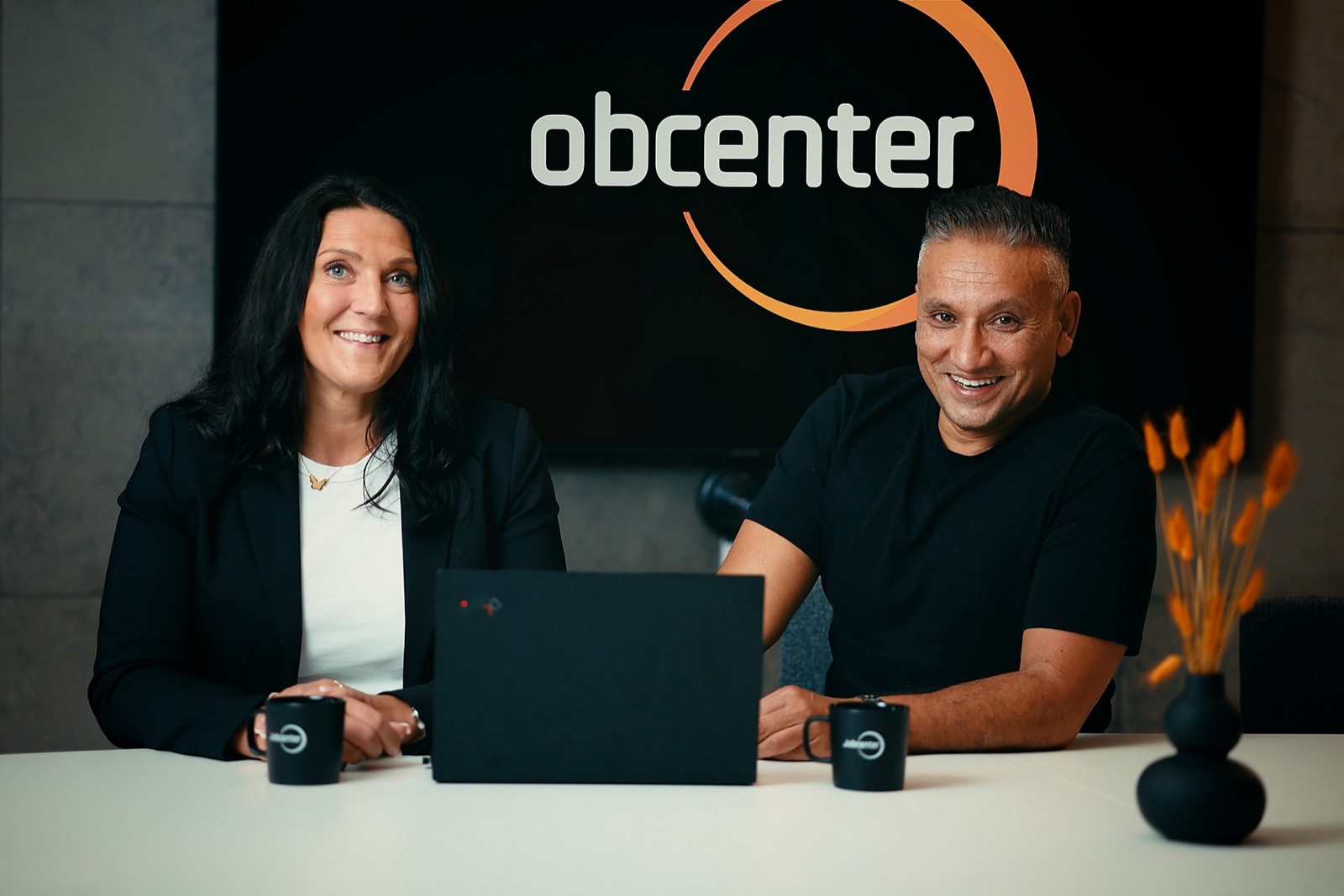 Två personer, en kvinna i kavaj och en man i svart t-shirt, sitter leende vid en laptop framför en skärm med Jobcenters logotyp.