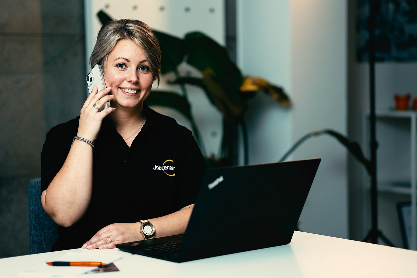 En kvinna i svart Jobcenter-piké pratar i mobiltelefon vid en laptop, med en grön växt i bakgrunden.