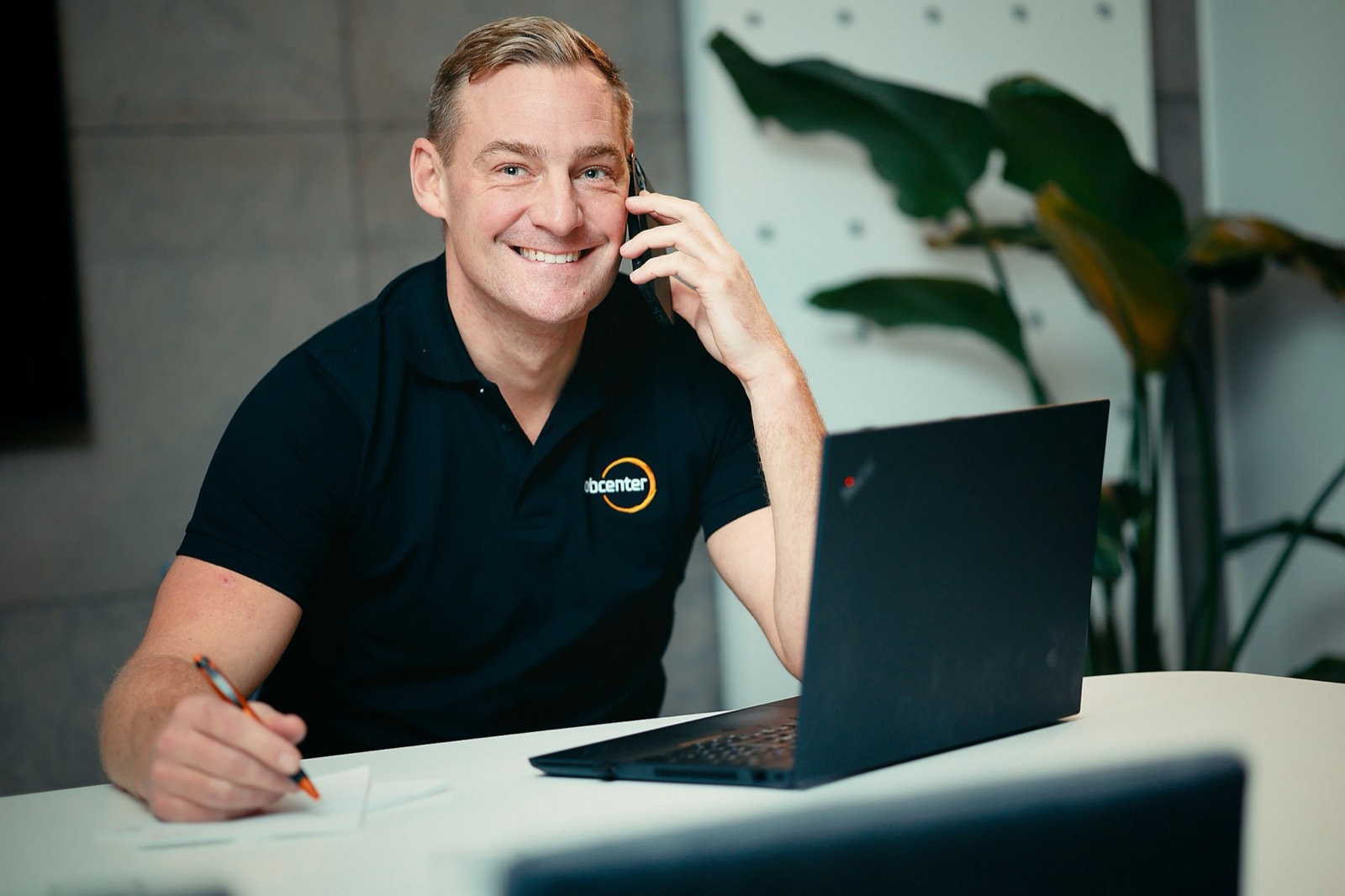 En man i svart Jobcenter-piké ler och pratar i mobiltelefon medan han antecknar vid sin laptop.