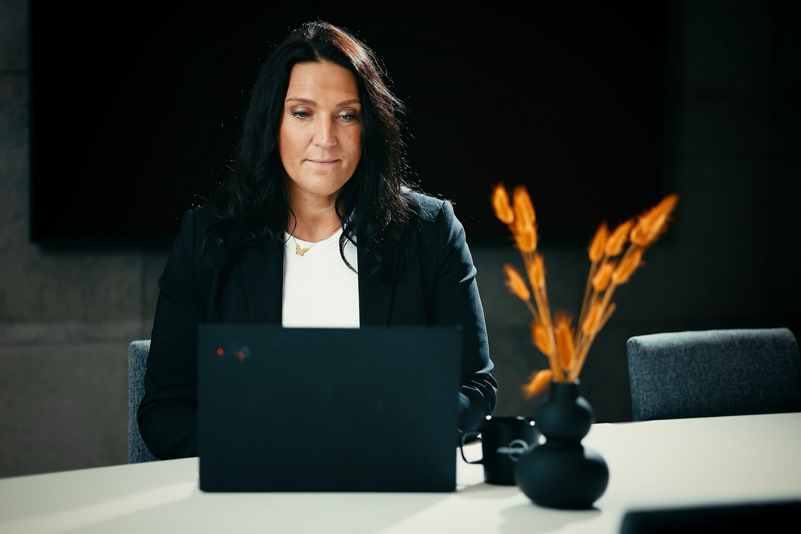 En kvinna i svart kavaj arbetar vid en laptop i ett mötesrum, bredvid en svart vas med orange torkade blommor.