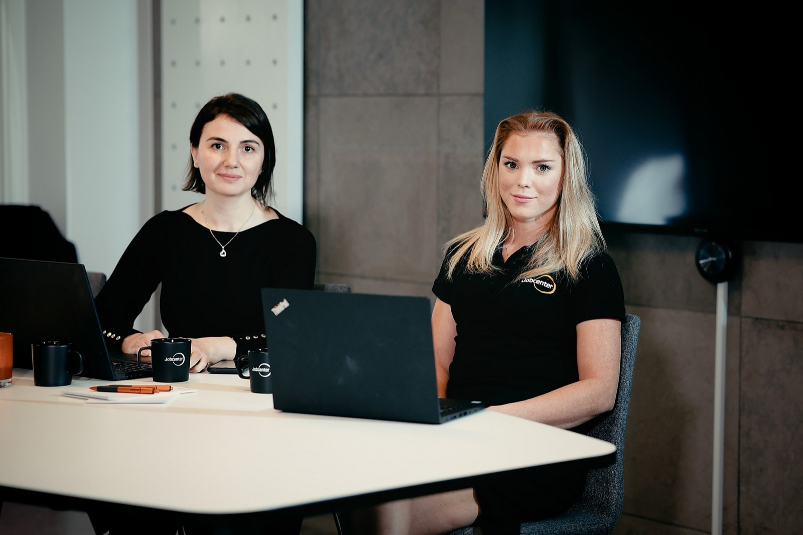 Två kvinnor sitter vid var sin laptop i ett mötesrum, med svarta Jobcenter-muggar på bordet.