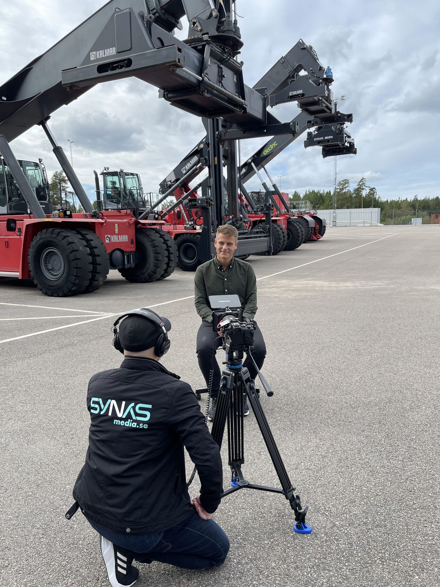 Outdoor interview in front of large forklifts at a company site — the film crew behind the camera