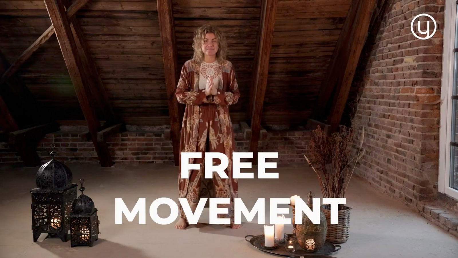 Stillbild ur en av utbildningsfilmerna: en kvinna i mönstrad dräkt står barfota med händerna samlade framför bröstet på en vind med träbjälkar, lyktor och tända ljus, med texten FREE MOVEMENT och Younitys logotyp.